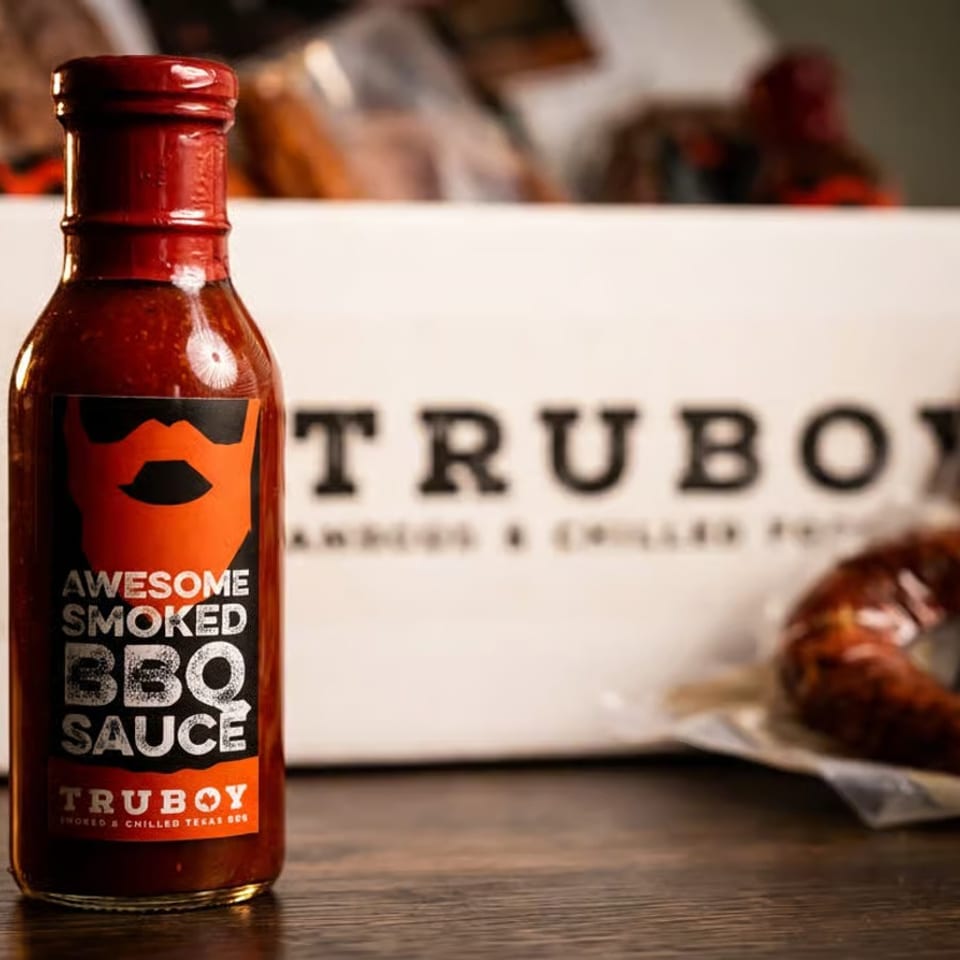 Bottle of Awesome Smoked BBQ Sauce displayed beside packaged Truboy smoked meats and branded packaging.