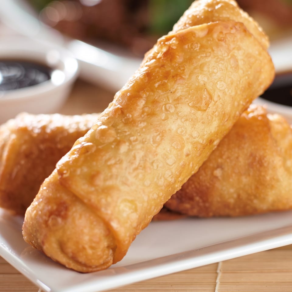 Golden fried triangular pastries arranged on a white plate over a wooden table, with blurred greens and dipping sauce in the background.