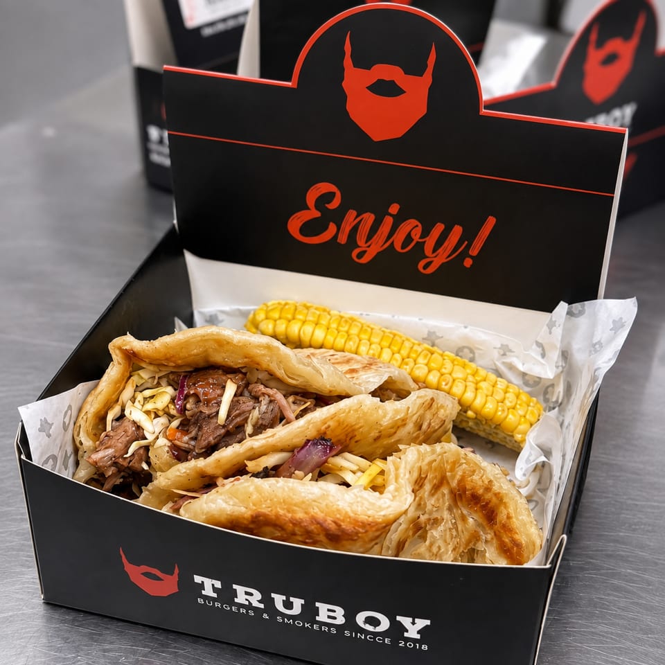 TruBoy takeout box filled with loaded flatbread sandwiches and grilled corn, featuring black-and-red branding.