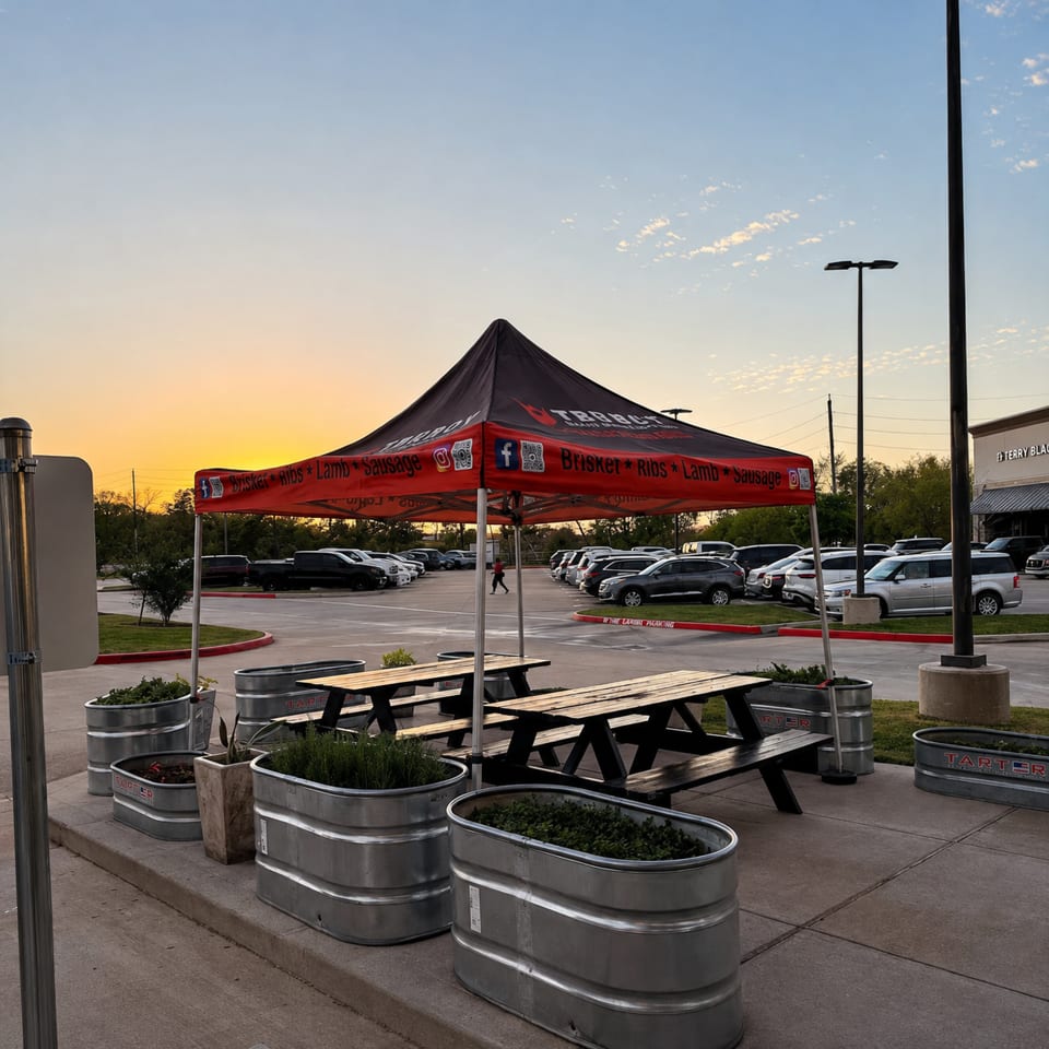 Outdoor barbecue restaurant seating with picnic tables, branded tent, metal planters, and parking lot at sunset.