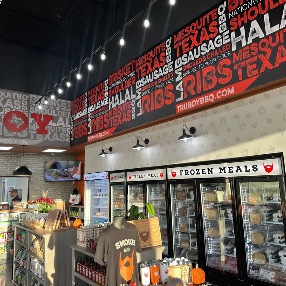 Tru-Bru BBQ restaurant interior with bold barbecue-themed wall graphics, frozen meal freezers, merchandise displays, and pendant lighting.