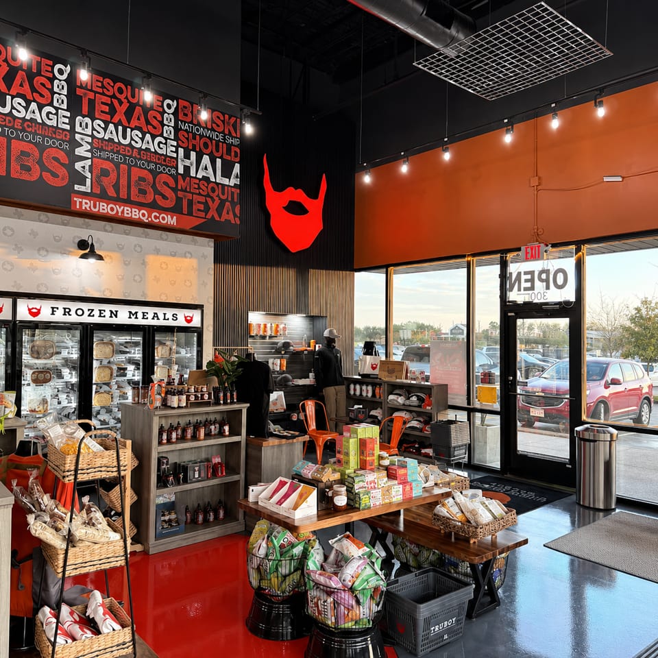 TruBuy BBQ restaurant interior featuring frozen meals, barbecue products, branded merchandise, red decor, and large windows.