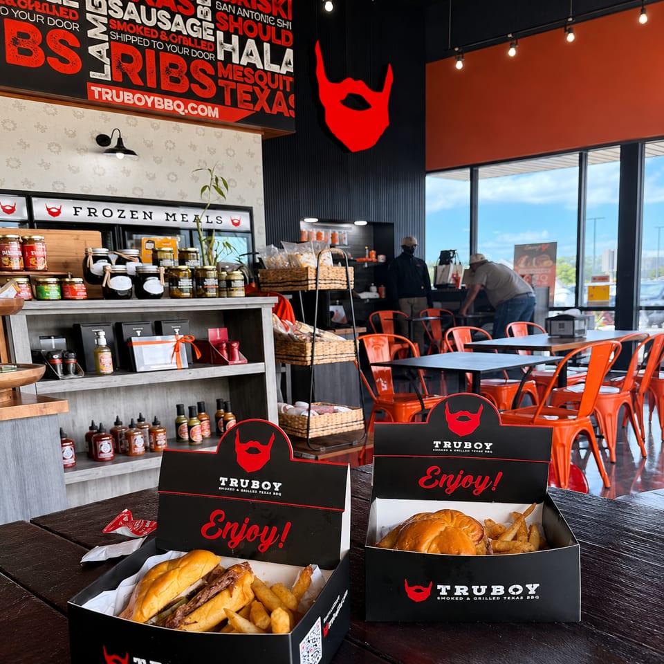 TruBoy BBQ restaurant interior with branded boxes of burgers and fries on a table, red chairs, sauces, and two diners.